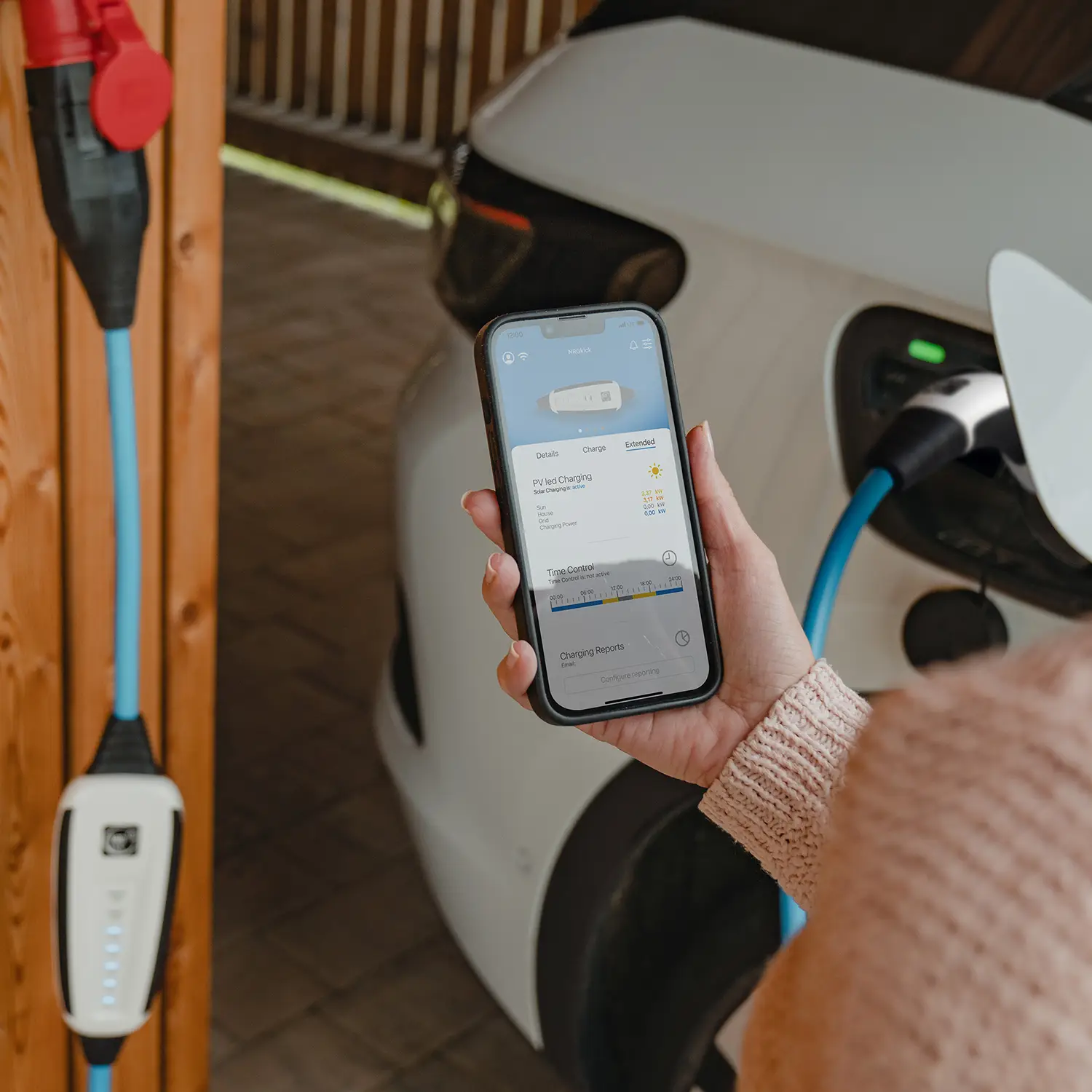 Electric car charges with NRGkick wallbox, woman demonstrates app on smartphone.
