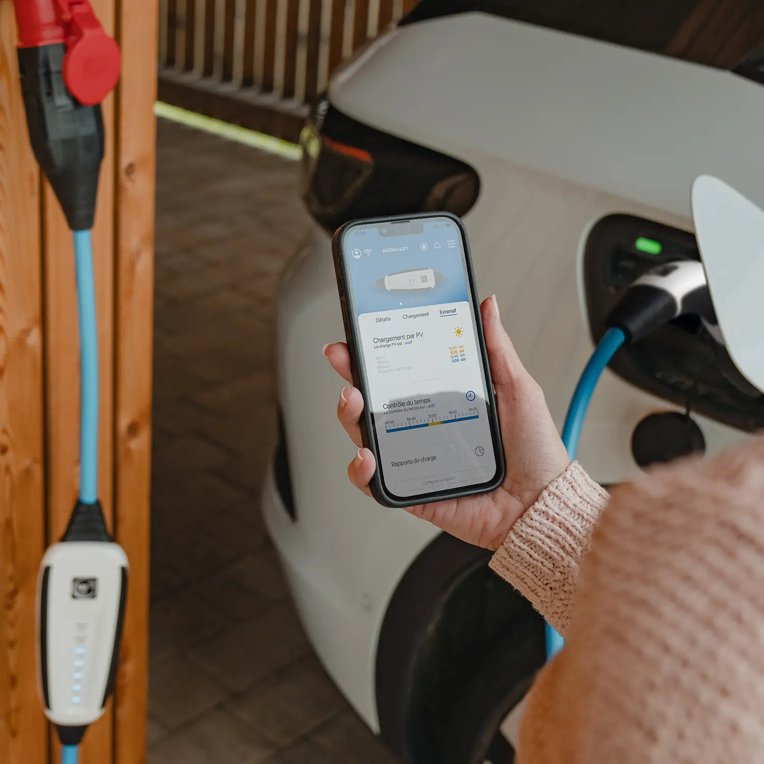 Une voiture électrique se recharge avec une Wallbox NRGkick, une femme fait la démonstration d'une application sur son smartphone.
